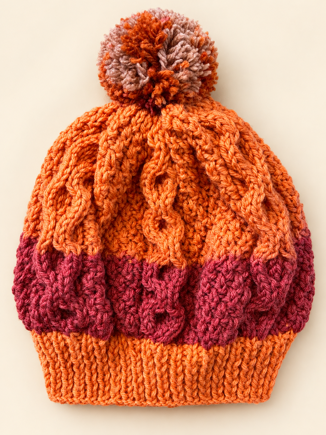 Overhead view of handmade Autumn Ember baby cable knit beret showing orange and burgundy cables, ribbed brim and pompom.