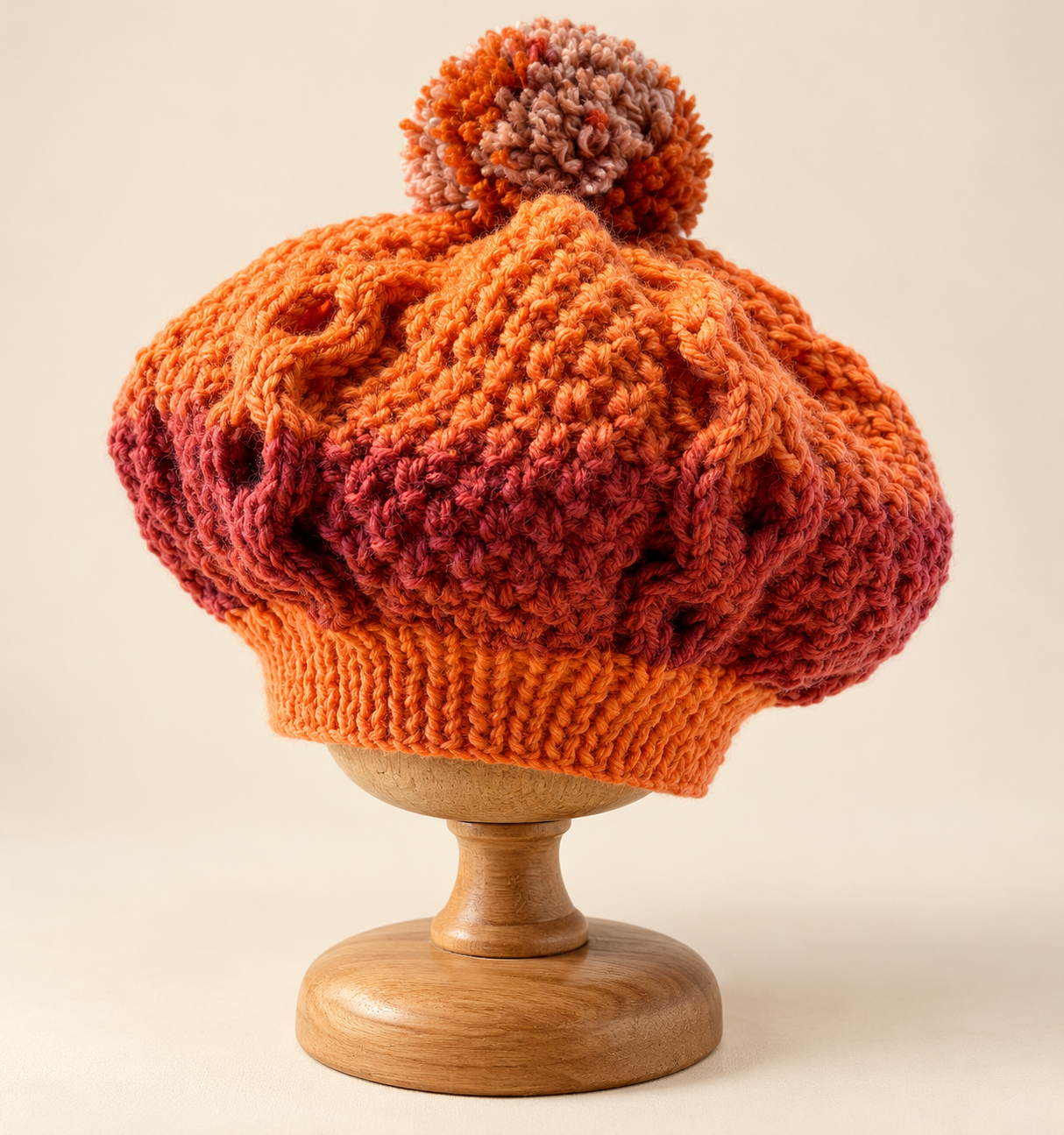 Hand knitted Autumn Ember baby cable knit beret in burnt orange and burgundy with multicoloured pompom, displayed on a wooden hat stand.