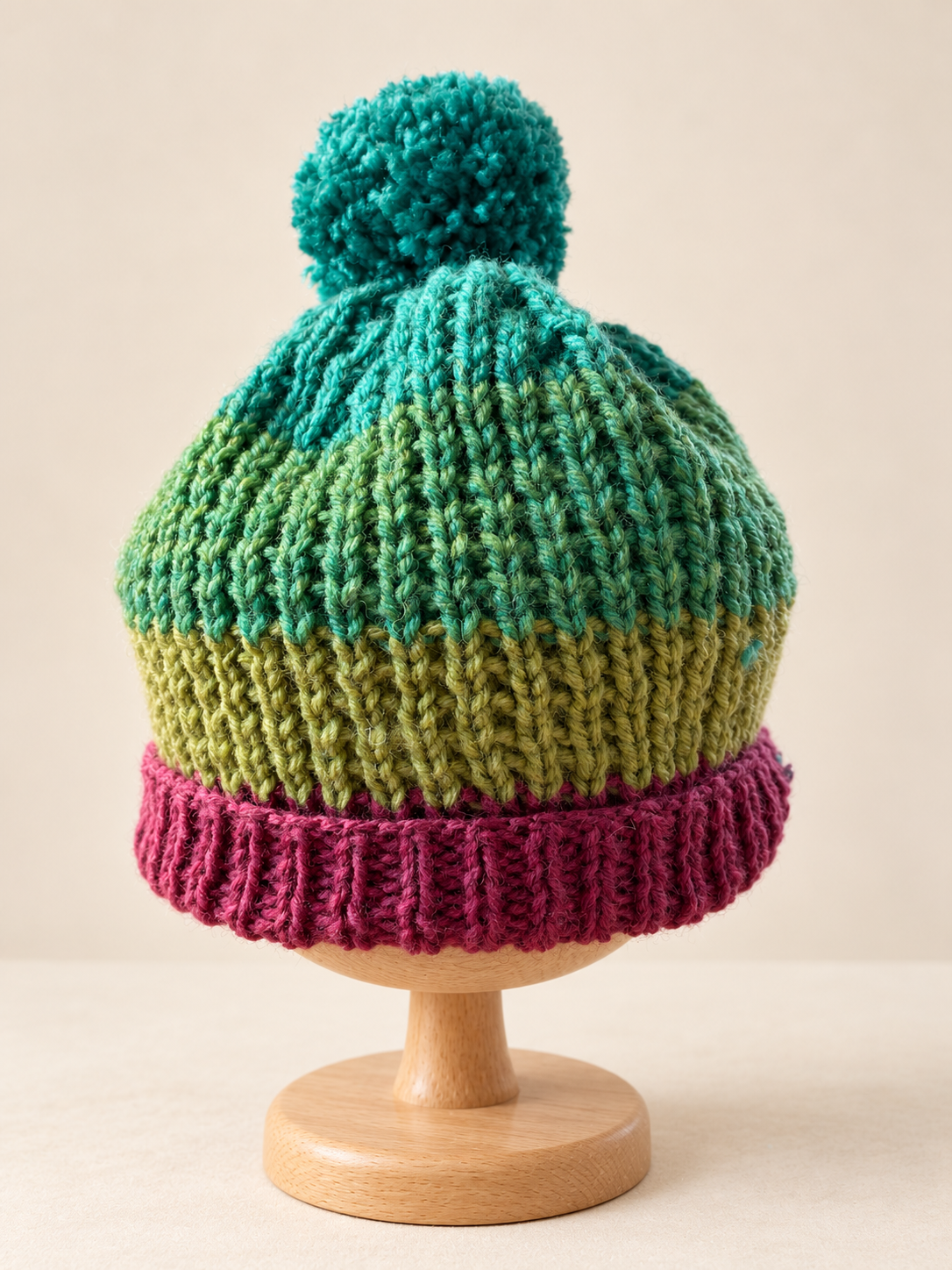 Teal and green Heather Cove pom-pom beanie displayed on a wooden stand, with a berry-purple folded brim